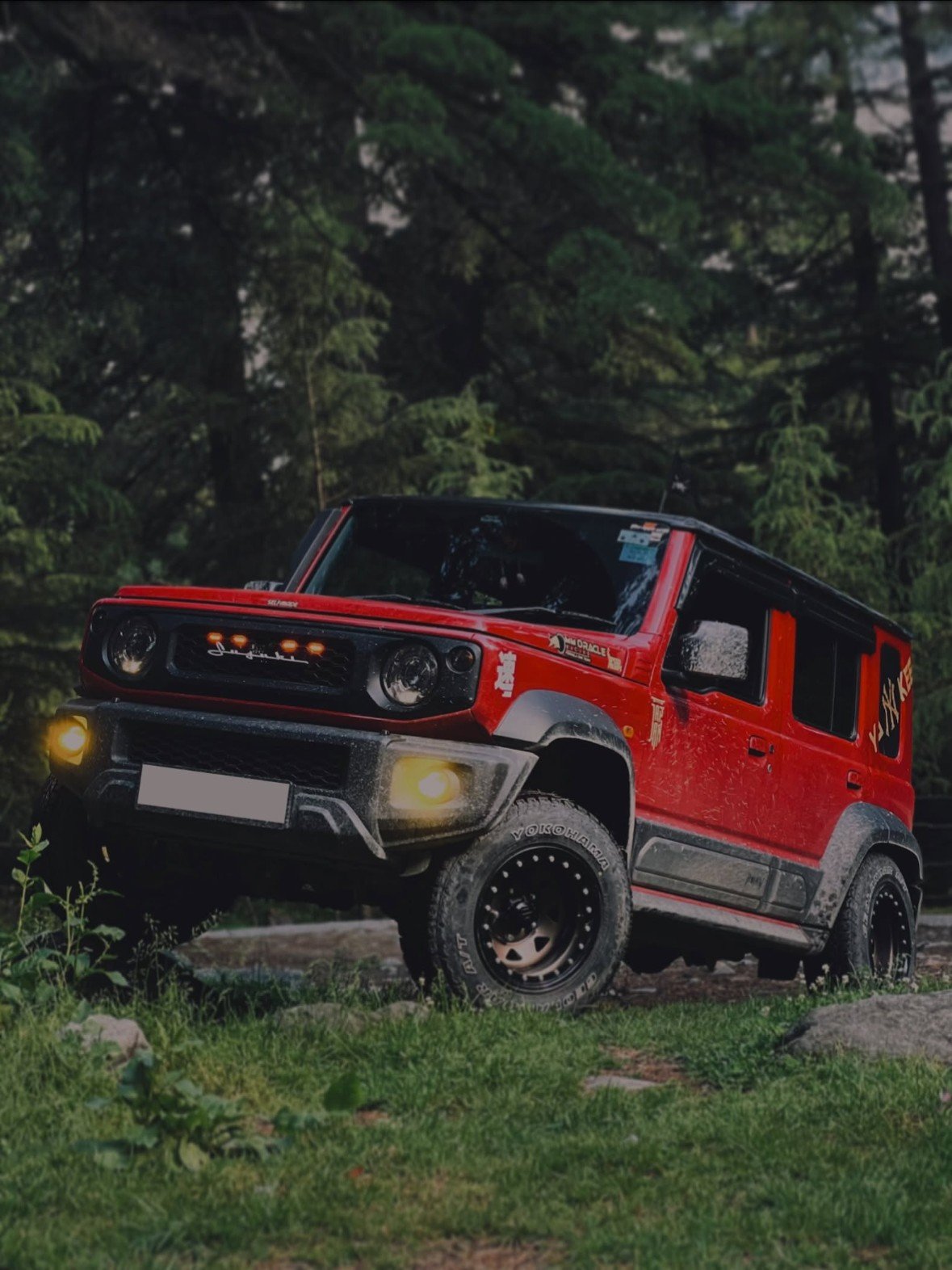 A red Maruti Suzuki Jimny with its fog lamps on, parked among pine trees