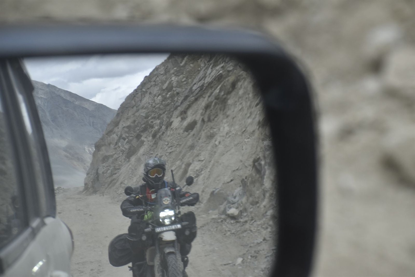 A motorcycle mirror reflecting a rider on the mountain road behind